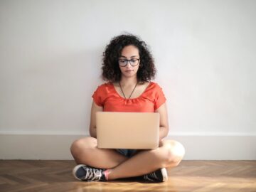 $24/hr – Project Coordinator (Bay Area, Remote) woman in orange crew neck t shirt sitting on brown wooden floor using laptop