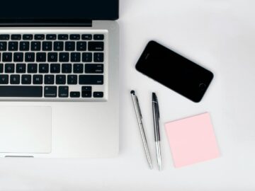 A top view of a modern workspace featuring a laptop, smartphone, pens, and a pink sticky note on a white desk.