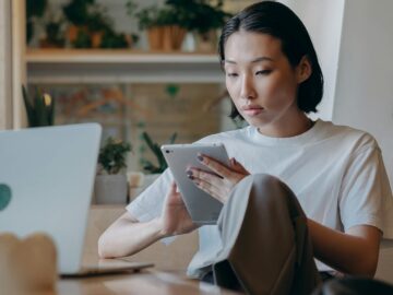 Focused Asian businesswoman using a tablet in a modern, plant-filled workspace with natural lighting.